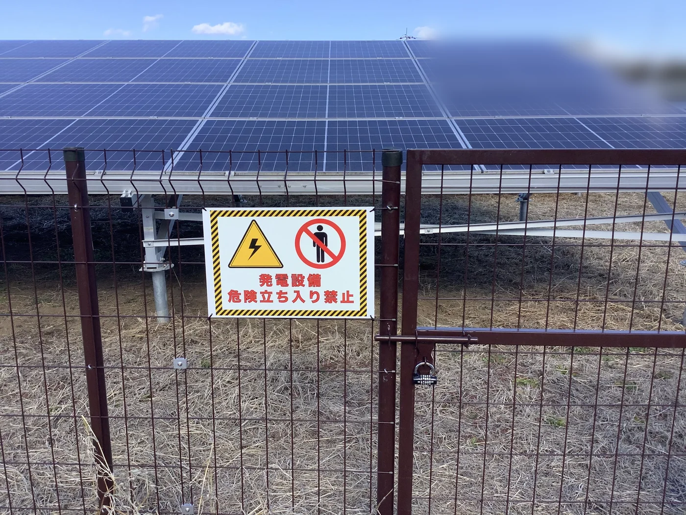 太陽光発電所のメッシュフェンスと扉。扉には南京錠が掛けられ、「発電設備 危険立ち入り禁止」の表示板が取り付けられている。奥に太陽光パネルが並ぶ。