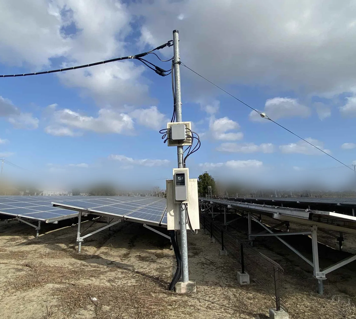 太陽光発電所の引込柱。柱に計器箱と接続箱が取り付けられ、足元から配管が地中へ伸びている。周囲には太陽光パネルの架台が並ぶ。